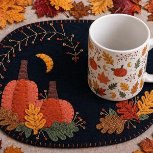 May include: A white ceramic mug with a fall-themed pattern of pumpkins, leaves, and moons sits on a navy blue felt placemat. The placemat features embroidered pumpkins, leaves, and a crescent moon design. Autumn leaves surround the placemat.