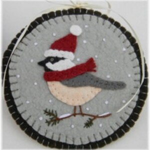 May include: A felt ornament depicting a gray and white bird wearing a red Santa hat and a red scarf, standing on a branch with green pine needles against a gray background with white snowflakes.