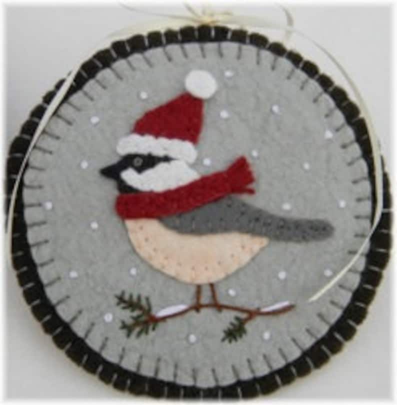 May include: A felt ornament depicting a gray and white bird wearing a red Santa hat and a red scarf, standing on a branch with green pine needles against a gray background with white snowflakes.