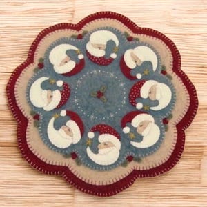May include: A round, felt Christmas placemat featuring a central design of Santa Claus faces in a circle. The placemat has a blue center, a beige border, and a scalloped burgundy edge. Small stars and holly accents are also included.
