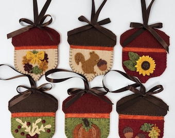 Puede incluir: Seis adornos de fieltro en forma de bellota con diferentes diseños de otoño, que incluyen una ardilla, un girasol, una calabaza y hojas. Cada adorno tiene una cinta marrón para colgar.