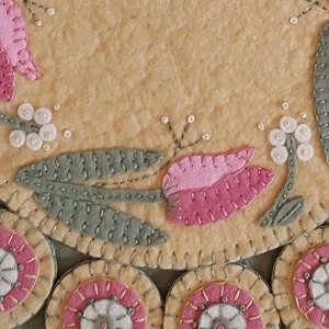 May include: A close-up of a handmade felt wall hanging with a floral design. The design features pink tulips, green leaves, and white beads. The background is a light beige color.