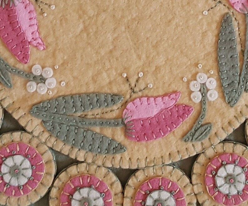 May include: A close-up of a handmade felt wall hanging with a floral design. The design features pink tulips, green leaves, and white beads. The background is a light beige color.
