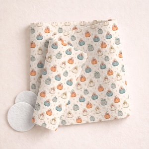 May include: A folded fabric with a repeating pattern of pumpkins in shades of orange, blue, and white, each adorned with a bow. The fabric is cream-colored with small floral accents. Three white, round pads are in the lower left corner.
