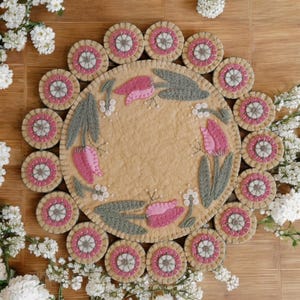 May include: A round wool rug with a floral design featuring pink tulips and green leaves. The rug is surrounded by a border of smaller circles with pink and white embroidery.