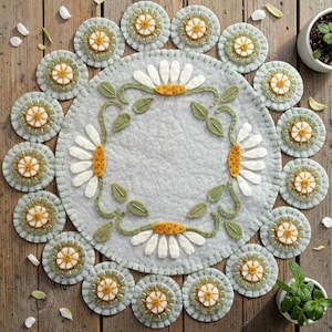May include: A light gray felt placemat and matching coasters with a daisy design. The placemat has a large daisy border, and the coasters have a smaller daisy design. The daisies have white petals and yellow centers. The placemat and coasters are on a wooden surface.