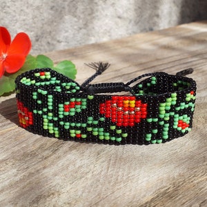 May include: Black beaded bracelet featuring a floral design in red, green, and yellow. The bracelet is adjustable with black cords. A red flower and green leaves are next to the bracelet.
