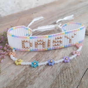 Puede incluir: Un brazalete de cuentas blancas con el nombre "DAISY" escrito en cuentas doradas. El brazalete tiene un borde de color rosa pastel, amarillo, azul y morado. Un segundo brazalete de cuentas blancas con abalorios de flores pastel se superpone.