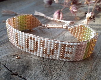 May include: A beaded bracelet with the word "LOVE" spelled out in gold and white beads. The bracelet is made with a pink, white, and green color scheme.
