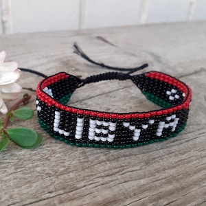 May include: A woven seed bead bracelet featuring the word LIBYA spelled out in white beads against a black background, bordered by red and green stripes. The bracelet includes black cord ties at the ends for an adjustable fit.