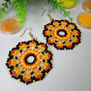 Puede incluir: Un par de pendientes de cuentas con un diseño circular. Los pendientes son de color negro, naranja, amarillo y crema. Los pendientes están hechos de cuentas pequeñas y tienen un patrón floral.