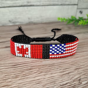 May include: A beaded bracelet featuring the flags of Canada and the United States. The bracelet is primarily red, white, blue, and black, with the flags' designs meticulously crafted with small beads. The adjustable closure is black.