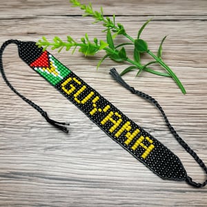 Könnte beinhalten: Ein schwarzes Perlenstirnband mit dem Wort "GUYANA" in gelben Perlen. Das Stirnband hat einen Abschnitt mit den Farben der guyanischen Flagge: rot, grün, gelb und schwarz. Das Stirnband hat an jedem Ende eine geflochtene schwarze Kordel.
