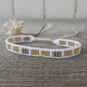 May include: White and gold beaded bracelet with a geometric pattern. The bracelet is made with small, square beads and has a simple, minimalist design.