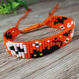 Peut inclure: Bracelet de perles orange avec des détails noirs, blancs et jaunes. Le bracelet présente un motif répétitif de formes noires et de perles blanches et jaunes sur un fond orange. Le bracelet est réglable.