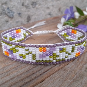 May include: A beaded bracelet featuring a floral design in shades of purple, green, orange, and white. The bracelet is tied with a white cord and has a small tassel.
