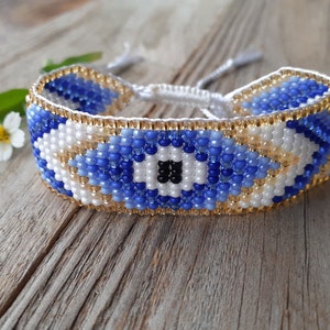 May include: A blue and white beaded bracelet with a gold trim and an evil eye design. The bracelet is made with small beads and has a tassel on each end.