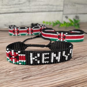 May include: Three beaded bracelets displaying the word "KENYA" in white beads against a black backdrop. The bracelets incorporate the Kenyan flag's colours: red, black, green, and white. They are presented on a wooden surface.