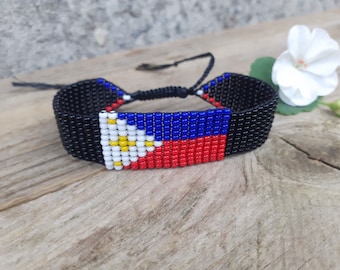 Puede incluir: Un brazalete de cuentas negras con una bandera de Filipinas tejida. La bandera es roja, blanca y azul con un sol amarillo en el centro.