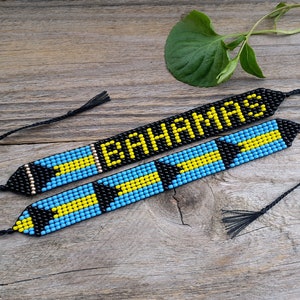Pode incluir: Duas pulseiras de contas com laços de corda preta. Uma pulseira tem a palavra "BAHAMAS" escrita em contas pretas. A outra pulseira tem as cores da bandeira das Bahamas em um padrão repetitivo.