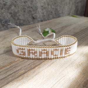Op de afbeelding: Witte kralenarmband met het woord "GEMINI" in gouden kralen. De armband heeft een gouden rand en een witte koordsluiting. De armband wordt gepresenteerd op een houten oppervlak.