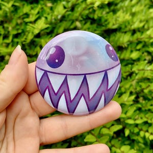 May include: A circular badge with a cartoon face design. The badge has a purple and white colour scheme with a galaxy-like background. The face has large purple eyes and a jagged, toothy grin.