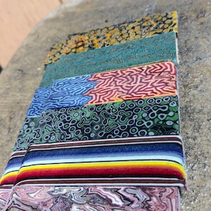 May include: A stack of colorful, patterned rectangular blocks. The blocks display various designs, including swirls, geometric shapes, and stripes in shades of pink, blue, green, and yellow. The blocks are stacked on a textured surface.
