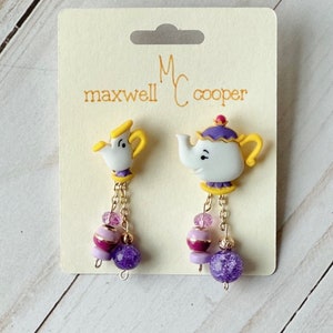 May include: A pair of dangle earrings featuring a teapot and a teacup charm with purple, pink, and gold accents. The earrings are labeled "Maxwell Cooper".