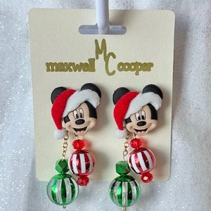 May include: A pair of Christmas-themed dangle earrings featuring Mickey Mouse wearing a Santa hat. The earrings have red and white striped beads, green and white striped beads, and red crystal beads.