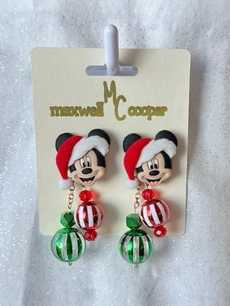 May include: A pair of Christmas-themed dangle earrings featuring Mickey Mouse wearing a Santa hat. The earrings have red and white striped beads, green and white striped beads, and red crystal beads.