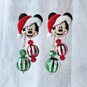 May include: A pair of Christmas-themed earrings featuring a Mickey Mouse character wearing a Santa hat. Each earring has a red and white striped ornament, a green and white striped ornament, and a gold chain.