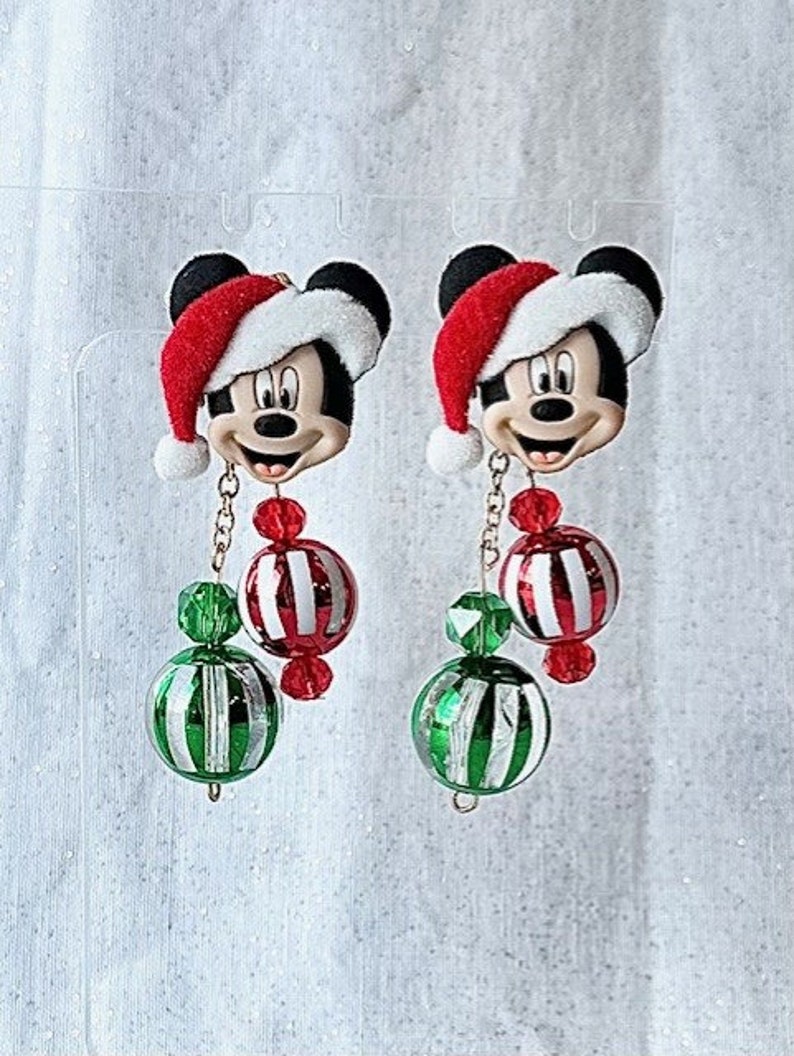 May include: A pair of Christmas-themed earrings featuring a Mickey Mouse character wearing a Santa hat. Each earring has a red and white striped ornament, a green and white striped ornament, and a gold chain.