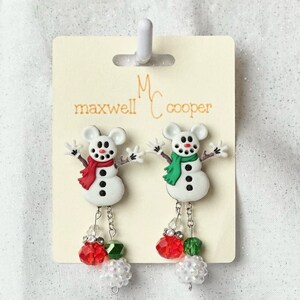 May include: A pair of snowman dangle earrings with red and green beads. The earrings are on a white card with the text "Maxwell Cooper" printed on it.