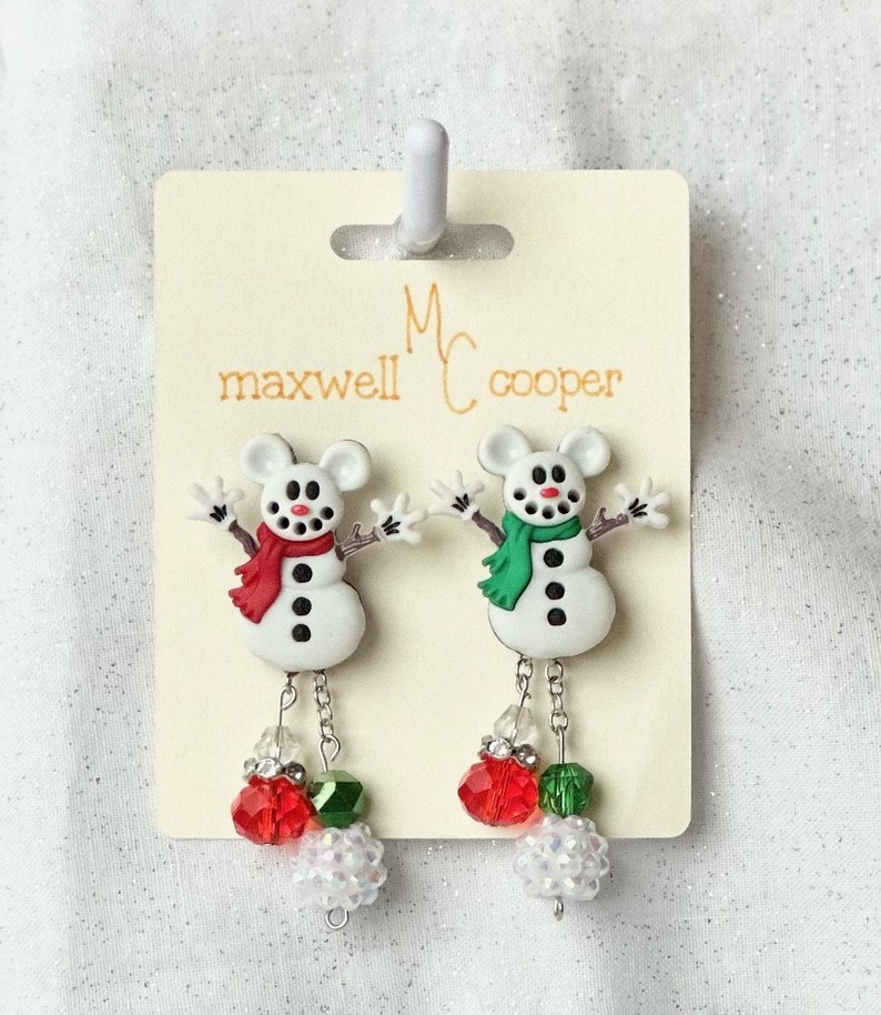 May include: A pair of snowman dangle earrings with red and green beads. The earrings are on a white card with the text "Maxwell Cooper" printed on it.