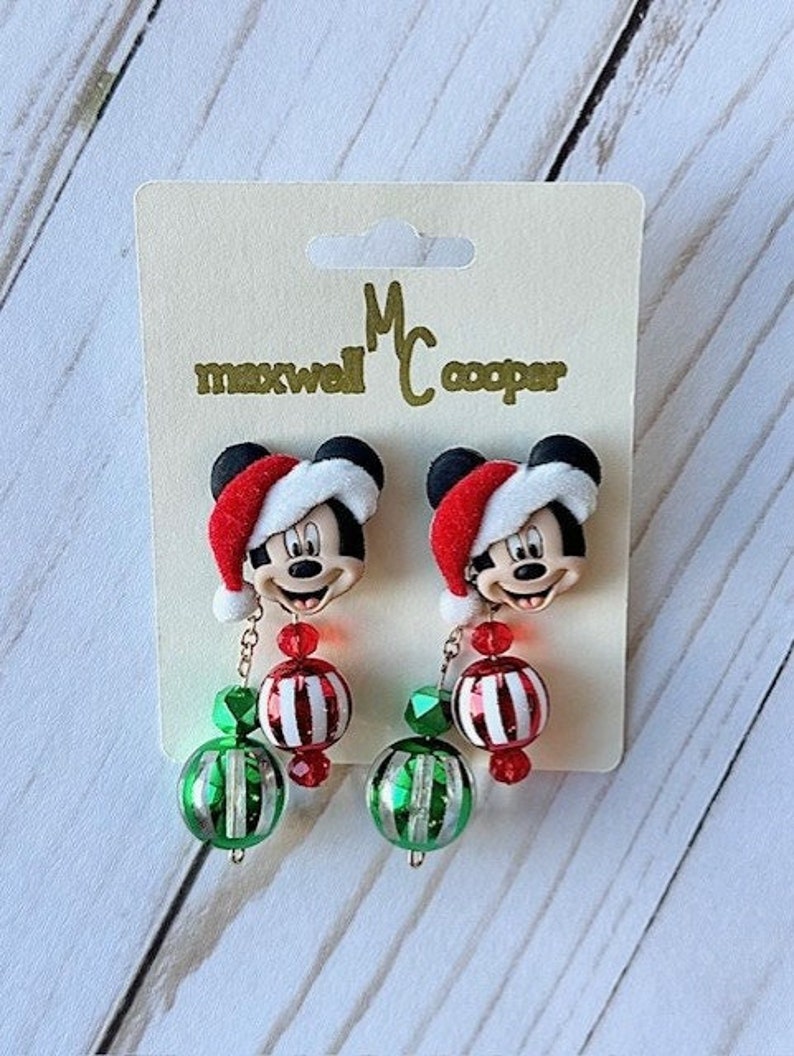 May include: A pair of Christmas-themed earrings featuring a Mickey Mouse character wearing a Santa hat. The earrings have red and white striped ornaments and green and silver beads.