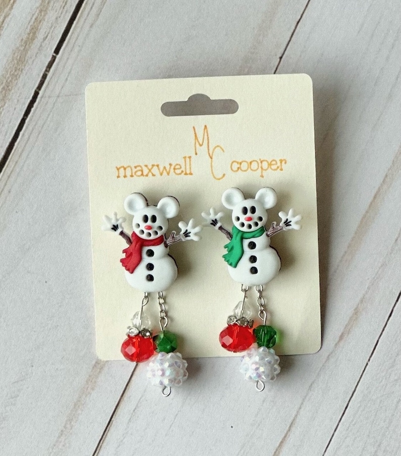 May include: A pair of snowman earrings with red and green accents. The earrings are made of polymer clay and feature a snowman with a red scarf and a green scarf. The earrings are hanging from silver chains and have red, green, and white beads.