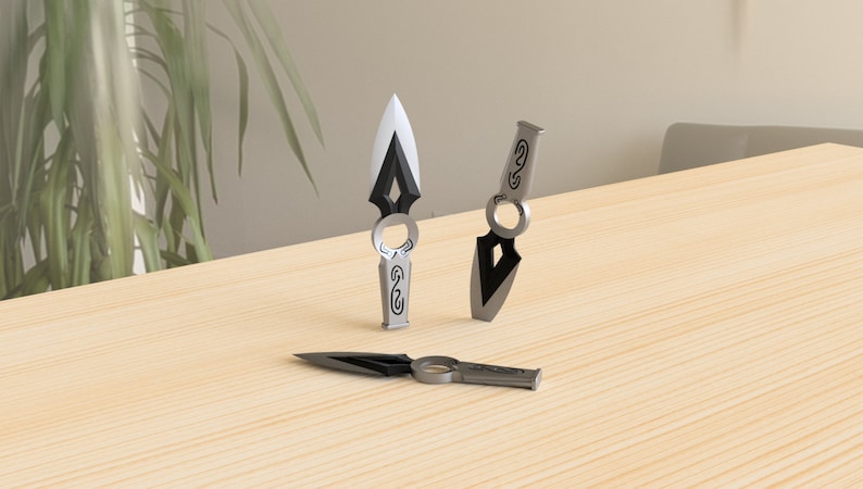 May include: Three silver throwing knives with black accents and intricate designs. The knives have a ring on the handle and a pointed tip.