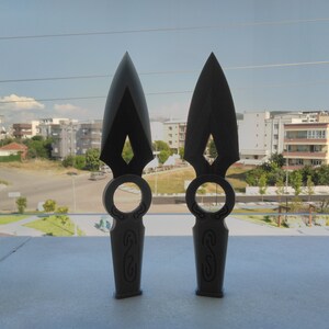 May include: Two black throwing knives with intricate designs on the handles. The knives have a sharp point and a ring on the handle.