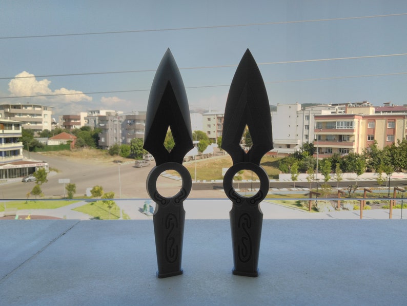 May include: Two black throwing knives with intricate designs on the handles. The knives have a sharp point and a ring on the handle.