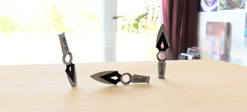 May include: Three silver throwing knives with black accents and intricate designs. The knives are on a light wood surface.