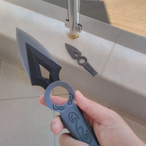 May include: Two black plastic throwing knives with intricate designs. The larger knife has a ring handle and the smaller knife has a pointed tip.