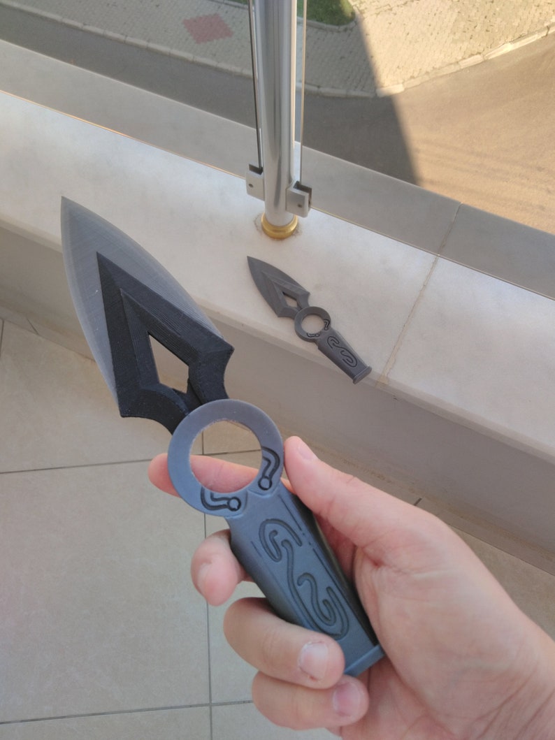 May include: Two black plastic throwing knives with intricate designs. The larger knife has a ring handle and the smaller knife has a pointed tip.