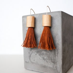 Könnte beinhalten: Ein Paar Holz-Quastenohrringe mit einer orangefarbenen Quaste und einem naturfarbenen Holzzylinder. Die Ohrringe werden auf einer grauen Betonfläche präsentiert.