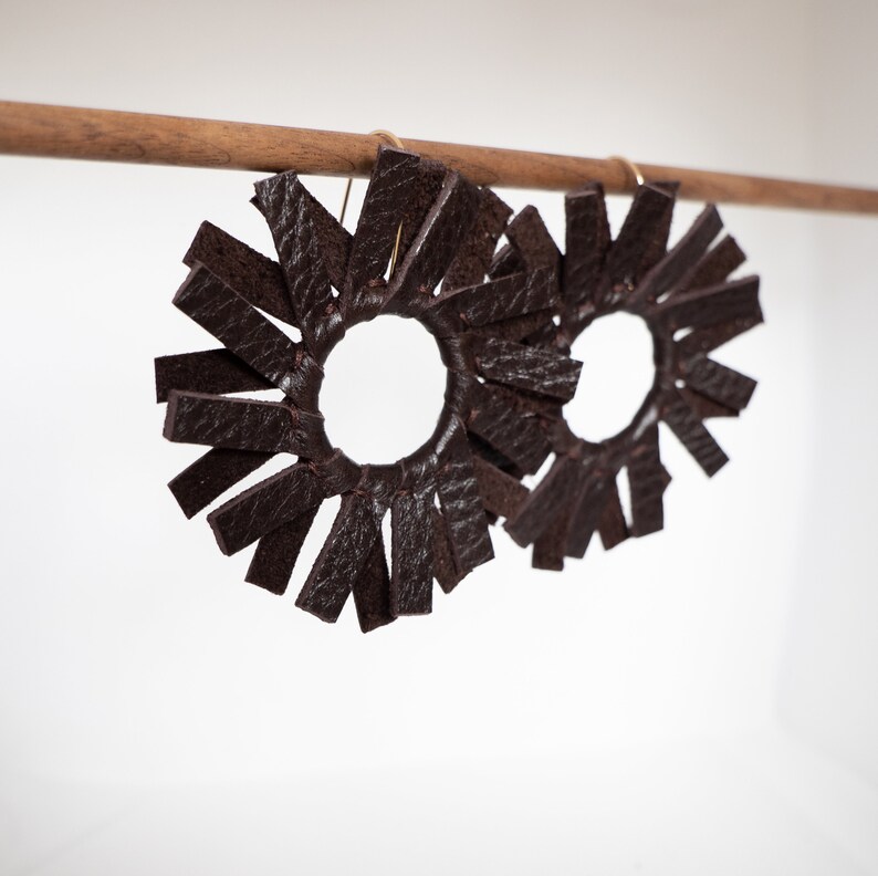 Angled shot focusing on the front of two dark brown espresso circular windmill shaped leather earrings hanging from a small walnut wood rod