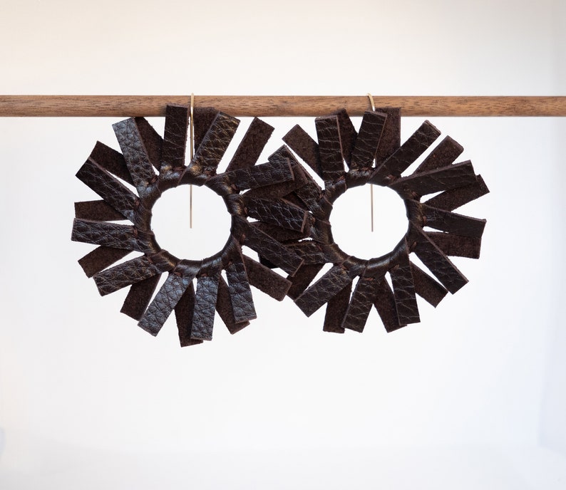 Pair of circular earrings hanging from a small walnut wood rod each made of dark brown espresso colored leather strips stitched together to create the look of a spinning windmill