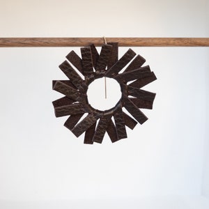 A single circular earring hanging from a small walnut wood rod made of dark brown espresso colored leather strips stitched together to create the look of a spinning windmill