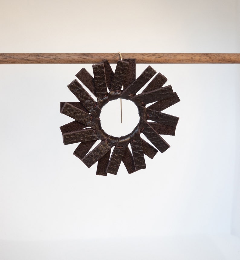 A single circular earring hanging from a small walnut wood rod made of dark brown espresso colored leather strips stitched together to create the look of a spinning windmill