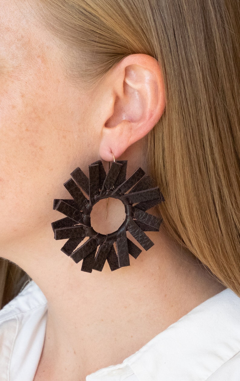 A circular dark brown espresso windmill shaped leather earring on the ear of a fair skinned model with straight dirty blonde hair.