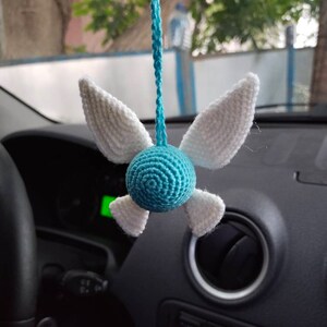 May include: A blue and white crocheted hanging ornament shaped like a character with wings. The ornament is hanging from a rearview mirror in a car.