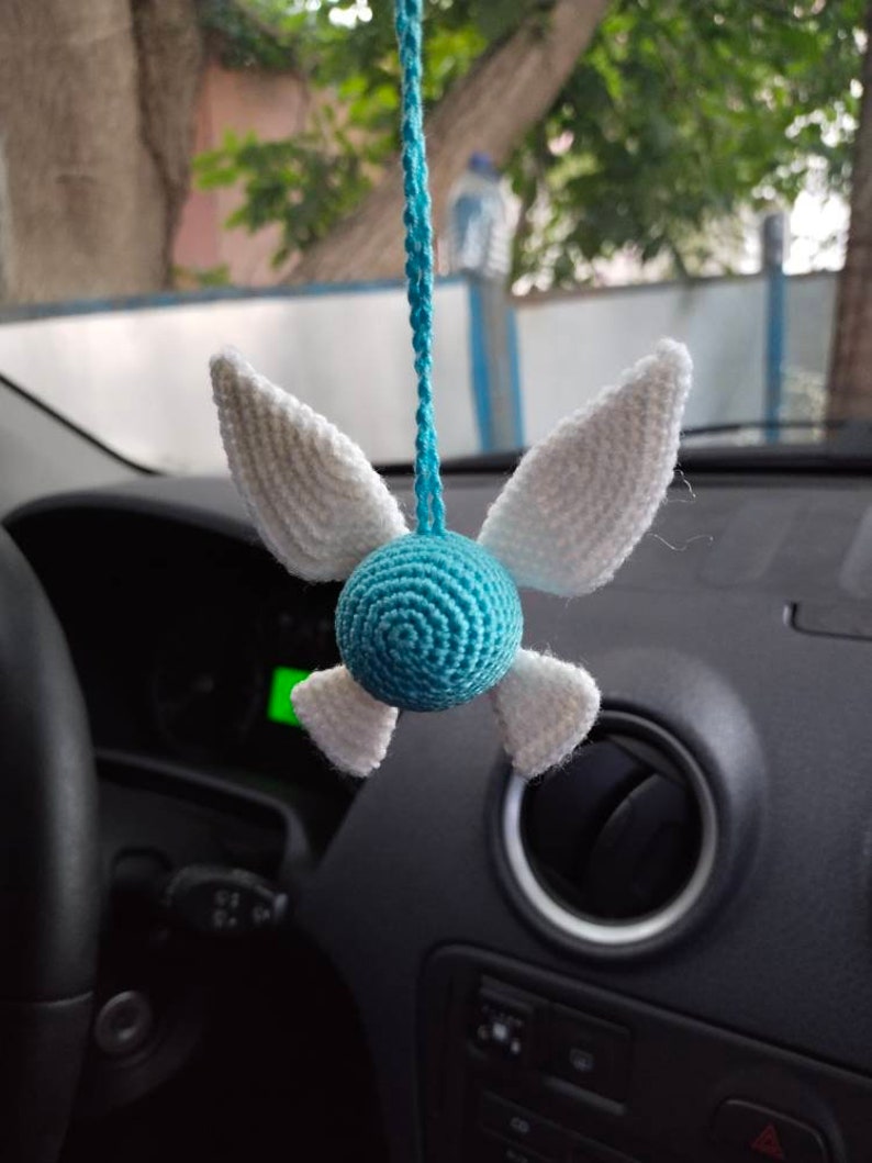 May include: A blue and white crocheted hanging ornament shaped like a character with wings. The ornament is hanging from a rearview mirror in a car.